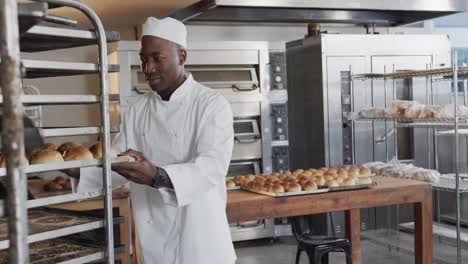 Portrait-of-happy-african-american-male-baker-in-bakery-kitchen-with-fresh-rolls-in-slow-motion