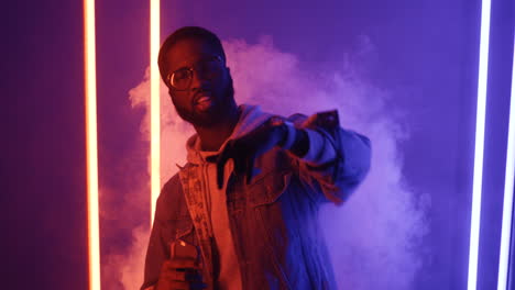 american stylish man in glasses dancing holding smartphone and looking at camera on pink and violet neon light background