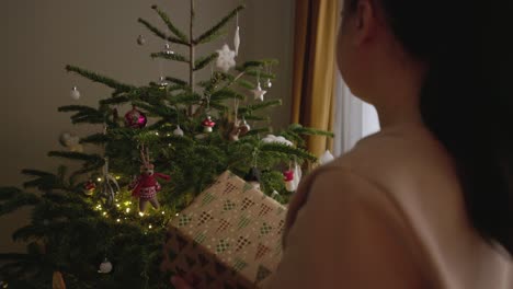 A-Girl,-Holding-a-Gift,-Examines-the-Decorations-Hanging-on-the-Christmas-Tree---Medium-Close-Up