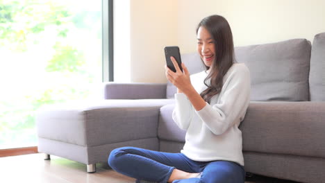 A-young-attractive-woman-sits-cross-legged-on-the-floor-while-she-visibly-and-happily-reacts-to-something-on-her-smartphone