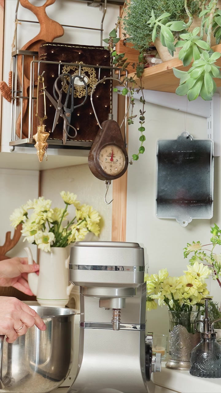 Girl places object onto household device. Holding and slightly moving metal bowl attached to mixer. Countertop scene includes metal rack with small objects wooden shelves
