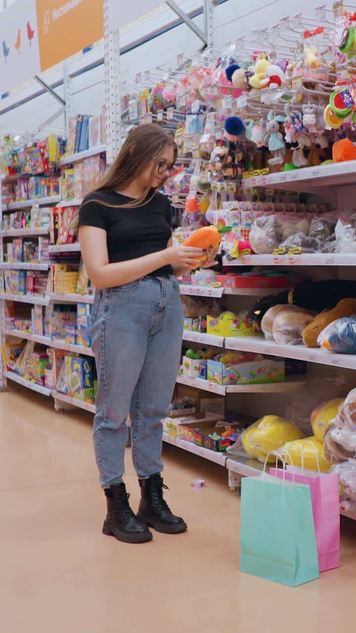 mujer con camiseta negra y vaqueros comprando en una tienda de juguetes, alcanzando un juguete de peluche suave en el estante, coloridos animales de relleno y juegos se alinean en los pasillos mientras las bolsas de compras pastel descansan en el suelo