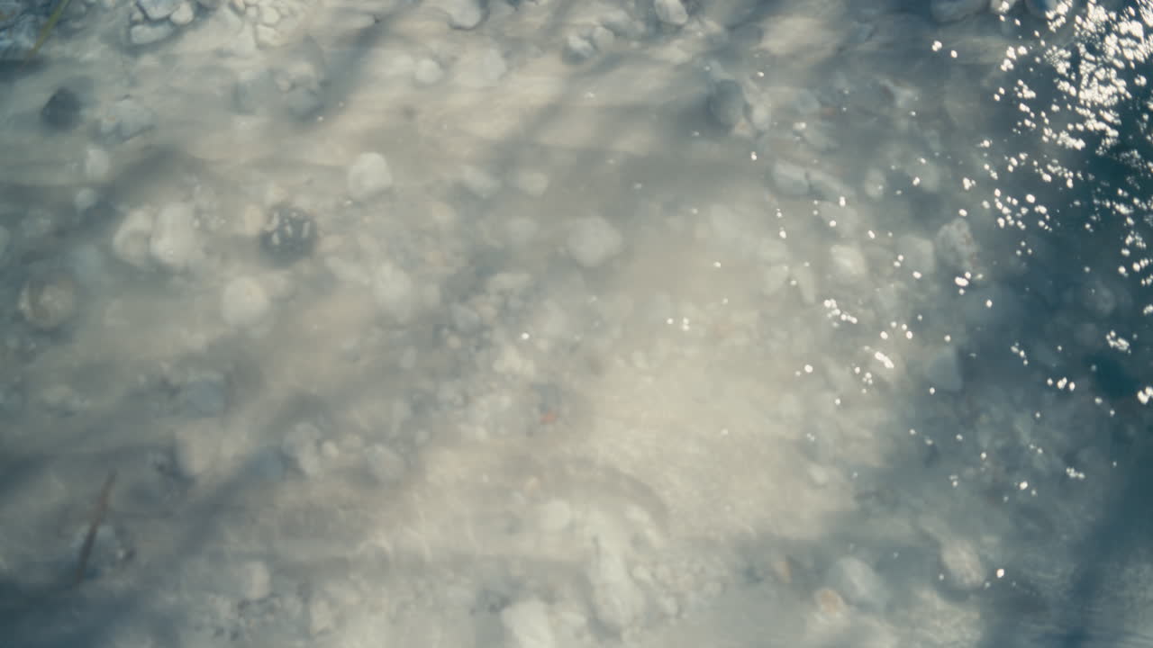 Vista astratta dall'alto di luce solare soffusa diffusa attraverso acqua limpida, che rivela pietre sotto la superficie. Lievi increspature e toni tenui creano uno sfondo naturale calmo e sognante