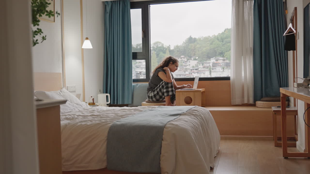 Hotel room writer types at window, solo guest writing on laptop on window seat, soft morning light, tidy bed with blue throw, city view beyond curtains, sipping tea between paragraphs, reflective