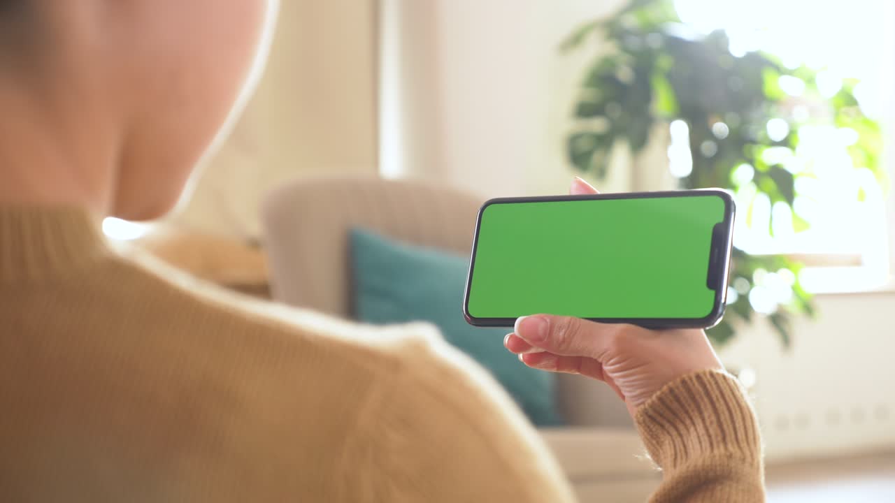 거실에서 녹색 스크린의 스마트폰을 사용하는 소녀 영화, 비디오 콘텐츠를 보고 있습니다. 소파에 누워있는 집에 있는 젊은 여성. 추적 마커. 수평 위치의 스마트 폰. 실내. 클로즈업