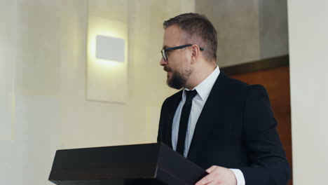 caucasian businessman speaker in formal clothes and eyeglasses on a podium at a conference, he is interacting with people in the hall during his speech