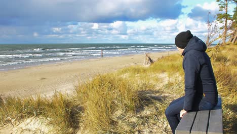 Caucasian-male-exploring-nordic-seaside-forest,-man-sitting-alone-on-the-gray-wooden-bench-on-the-beach,-coastal-pine-forest,-white-sand-beach,-healthy-activity-concept,-wide-shot