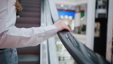 close-up of a woman hand resting on an escalator rail as she moves upwards in a modern shopping mall, she wears a white shirt and denim jeans with a phone visible in her back pocket