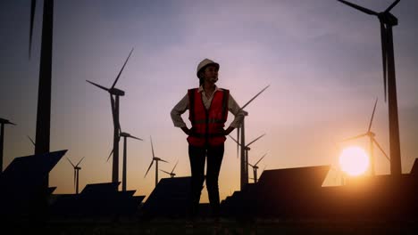 full body of asian female engineer in a helmet looking around while standing with arms akimbo in front of wind turbines rotating at sunset