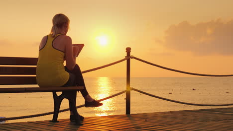 fitness woman enjoys a tablet sits on a bench on a pier against the backdrop of the rising sun 4k vi