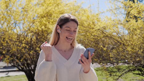 feliz mujer bonita en un suéter blanco usando teléfono móvil feliz mujer rubia celebrar victoria en línea, éxito escuchar grandes noticias sentirse asombrado apuestas deportivas en el parque cerca de un árbol con hojas amarillas.