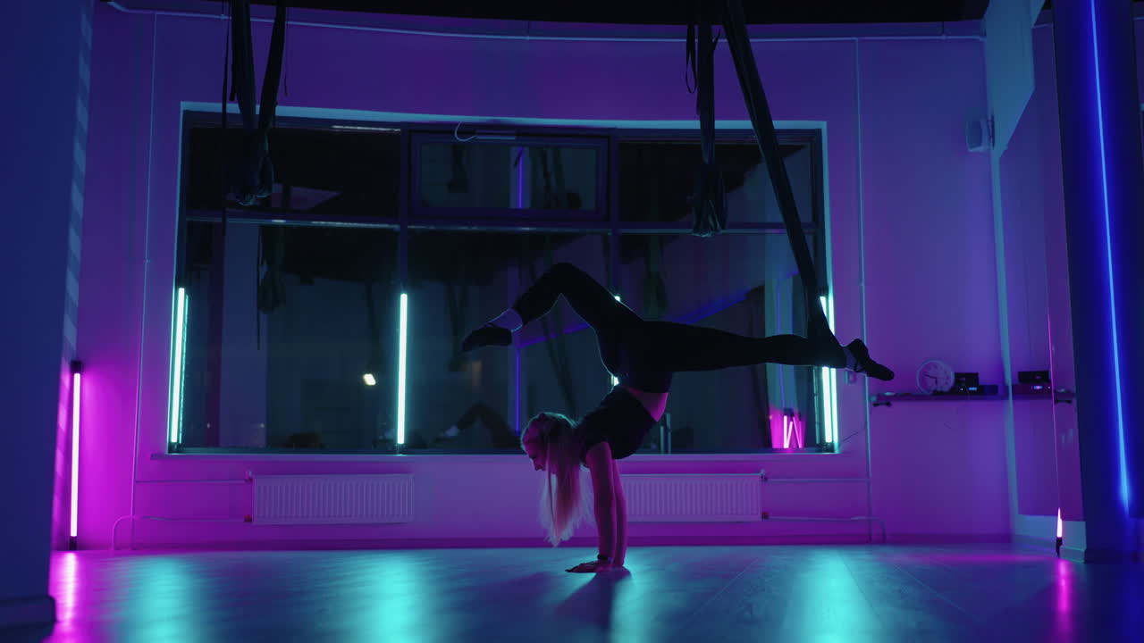 A woman does sports yoga in the air on a hammock in neon light performing graceful exercises and movements from stretching and dancing in slow motion.