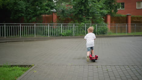 a young child rides a red scooter joyfully down a paved pathway bordered by green grass, metal railings, and a nearby building