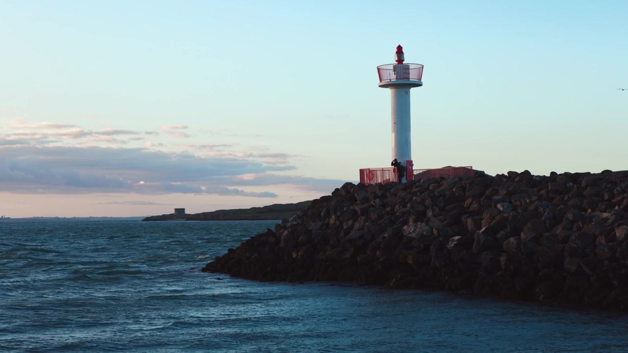Pasangan berdiri di samping Mercusuar Howth pada malam yang berangin dengan pemandangan Ireland’s Eye dan menara Martello