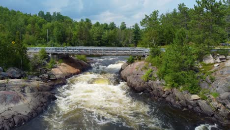 Stromschnellen-Des-Flusses,-Der-Unter-Der-Fußgängerbrücke-Mit-Dichtem-Wald-Fließt