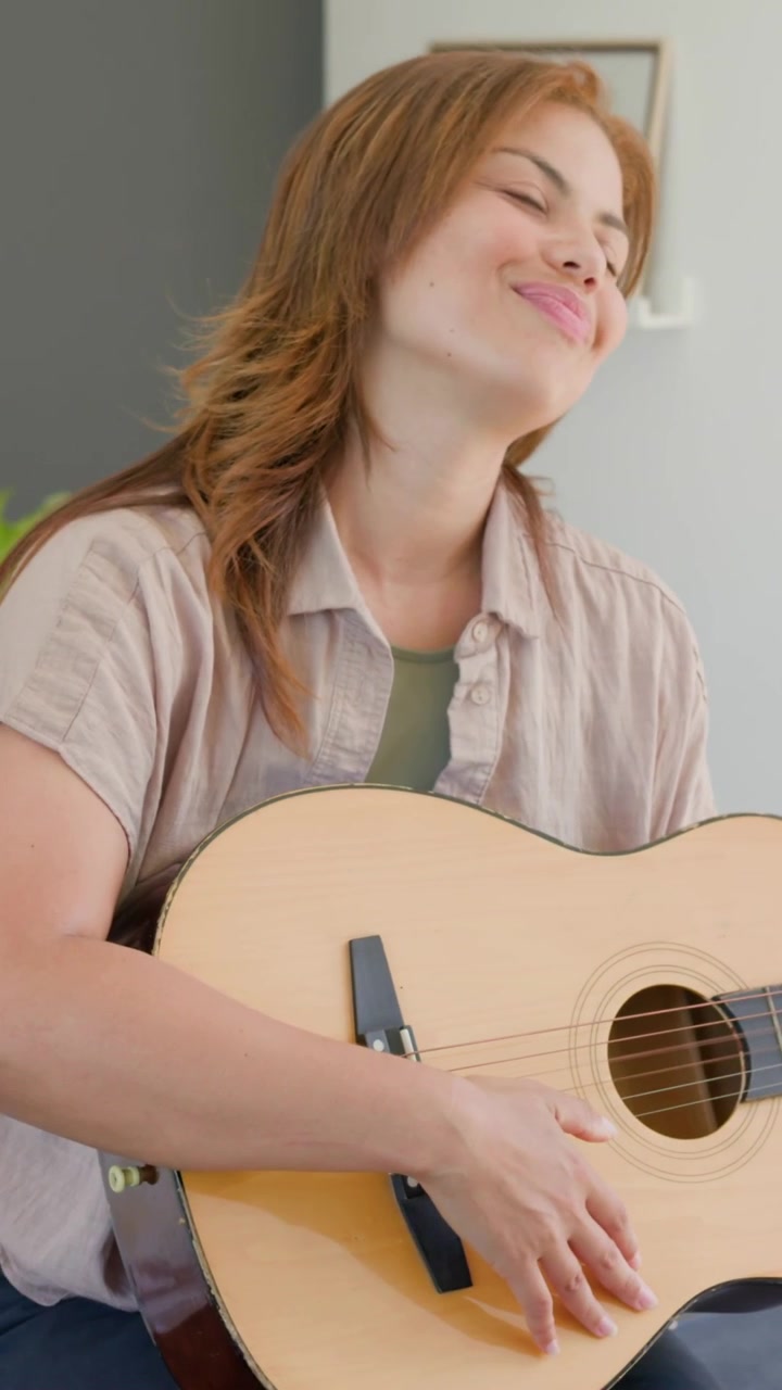 Video verticale: Posizionamento, donna che strimpella la chitarra, si esercita a casa, indossando una camicia beige, orologio.
