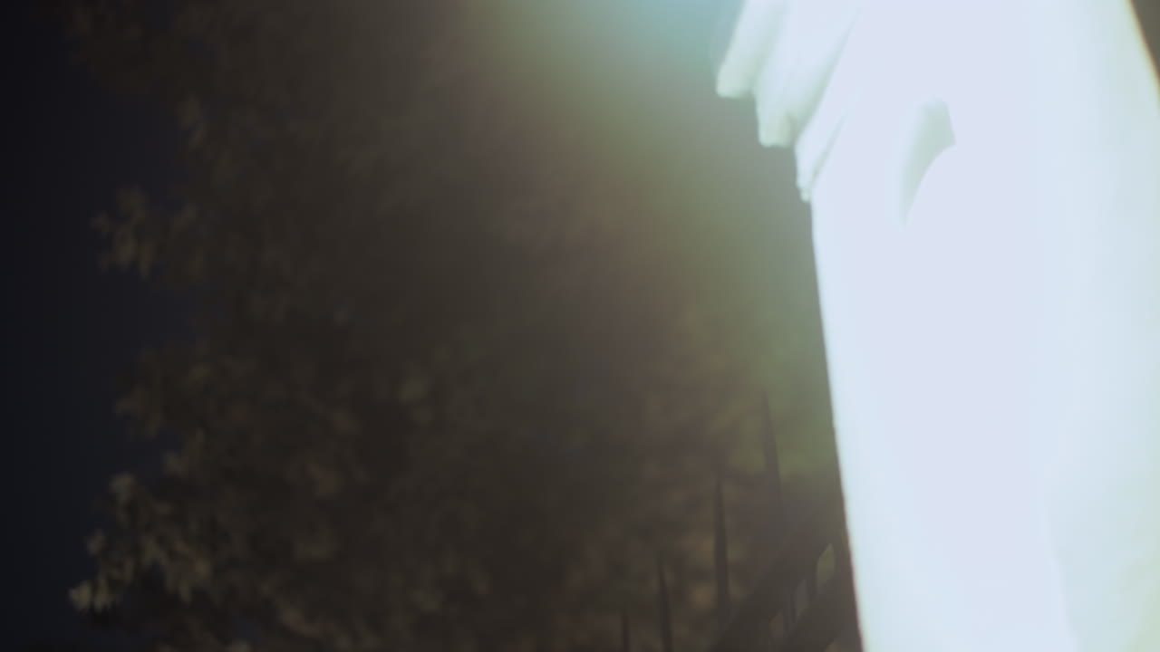 Close-up night view of glowing decorative iron fence illuminated by bright light from white pillar against dark blurred background, creating atmospheric urban scene with ambient glow