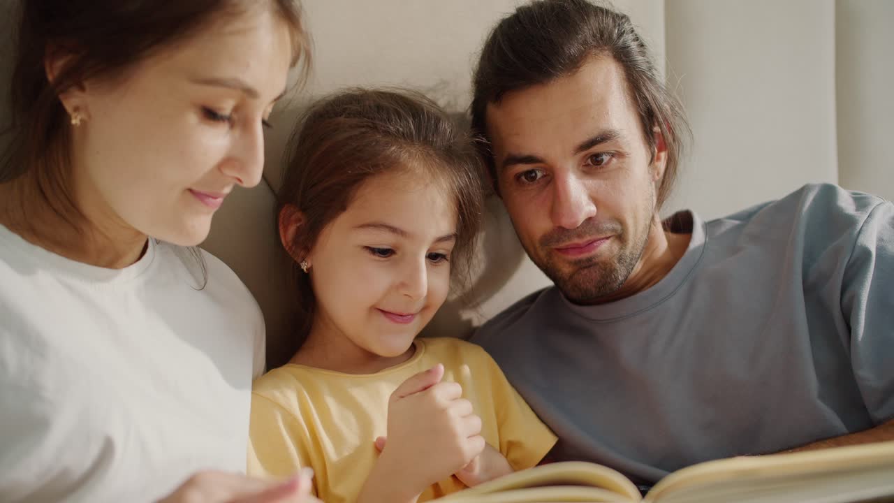 A little brunette girl with a yellow T-shirt is reading a book and looking at the pictures in it with her parents. A man and a woman are leafing through a book with their daughter