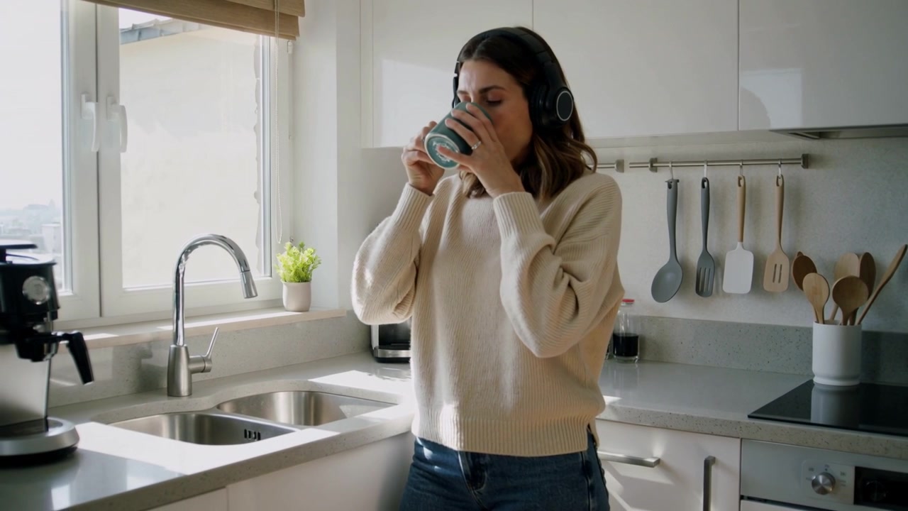 Donna in maglione e cuffie che, dopo averla tenuta, solleva una tazza verde, sorseggia e guarda fuori dalla finestra della cucina.