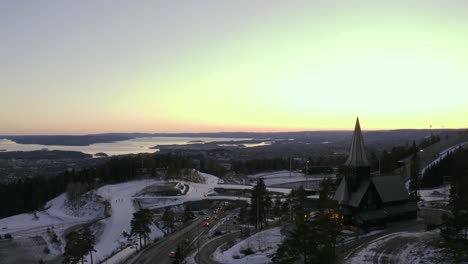 Oslo-city-drone-wrap-around-Holmenkollen-chapel-church,-with-Holmenkollbakken-ski-jump,-Oslo-Vinterpark-Winterpark-Tryvann-at-Sunset