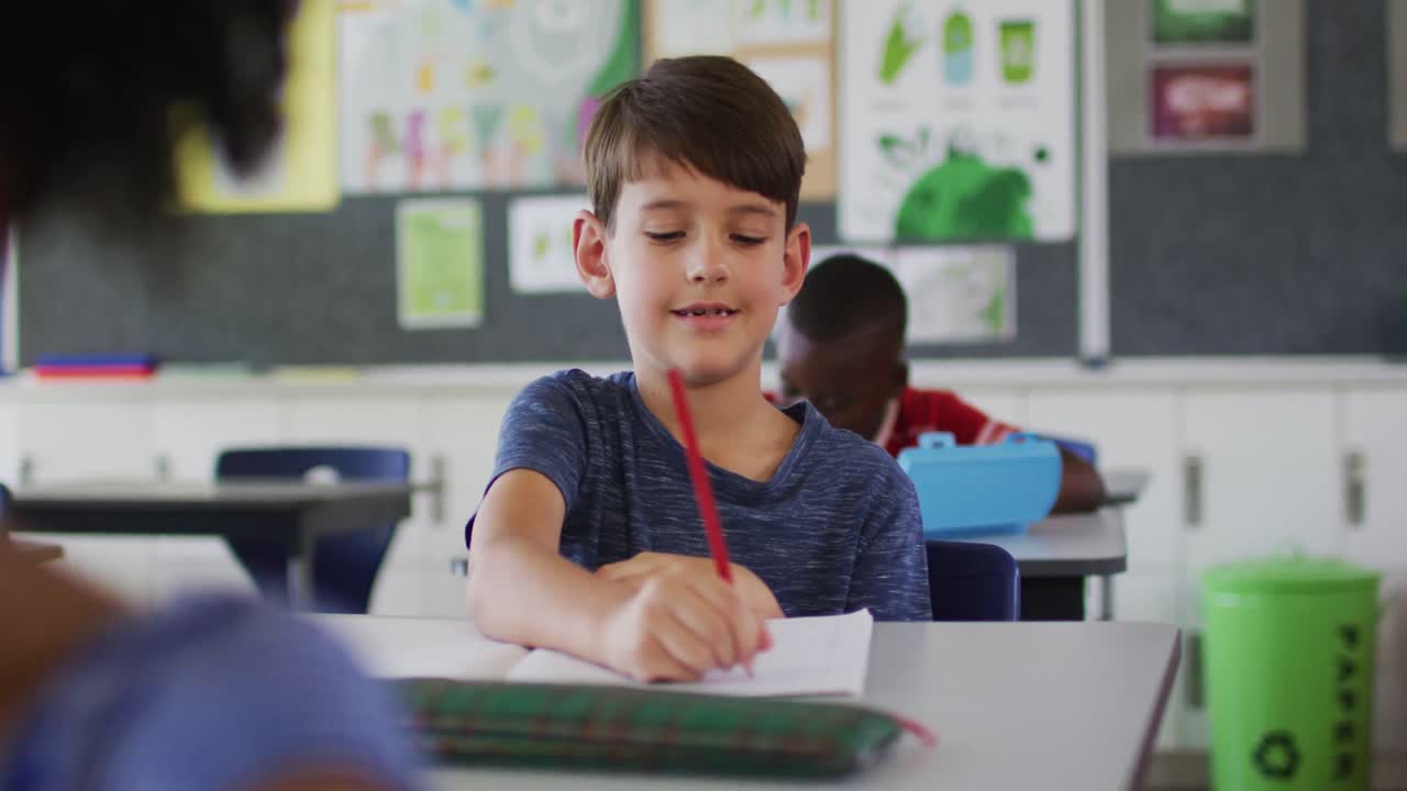 retrato de un niño de escuela caucásico feliz sentado en el aula, tomando notas, mirando a la cámara