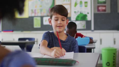 Portrait-of-happy-caucasian-schoolboy-sitting-at-classroom,-making-notes,-looking-at-camera