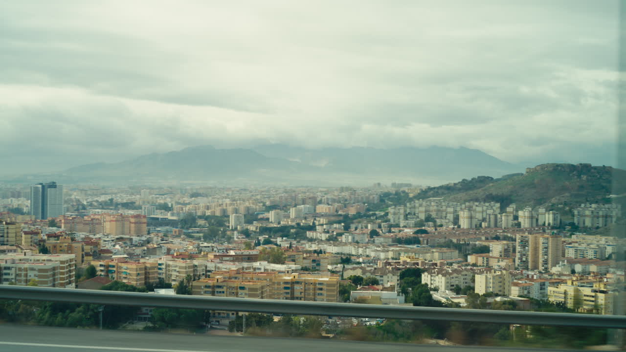Vista elevada de Málaga, España, filmada a través de la ventanilla de un coche, revelando densos edificios urbanos, colinas onduladas y montañas distantes bajo un cielo nublado