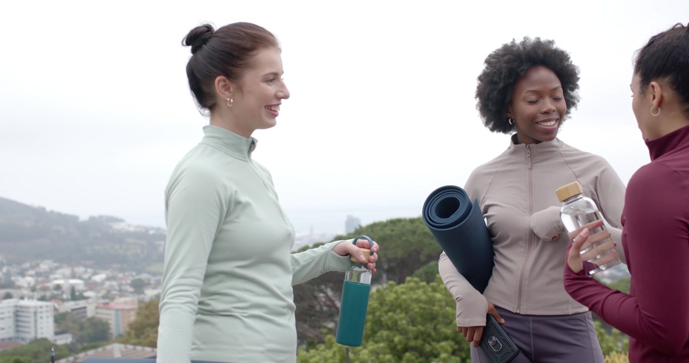 Tre donne in leggings che si salutano e parlano su una collina con tappetini e bottiglie, preparandosi per l'allenamento