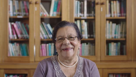 Porträt-Einer-Eleganten-älteren-Frau-Mit-Gemischter-Abstammung,-Die-Vor-Dem-Hintergrund-Einer-Bibliothek-Fröhlich-In-Die-Kamera-Lacht-Und-Eine-Perlenkette-Trägt