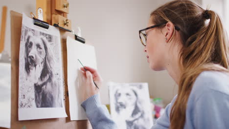 Female-Teenage-Artist-Sitting-At-Easel-Drawing-Picture-Of-Dog-From-Photograph-In-Charcoal