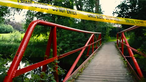 low angle footage of a yellow police crime scene do not enter tape pulled in front of a red metallic bridge with a boarded wooden pathway or road during a summer cloudy day with threes on the sides