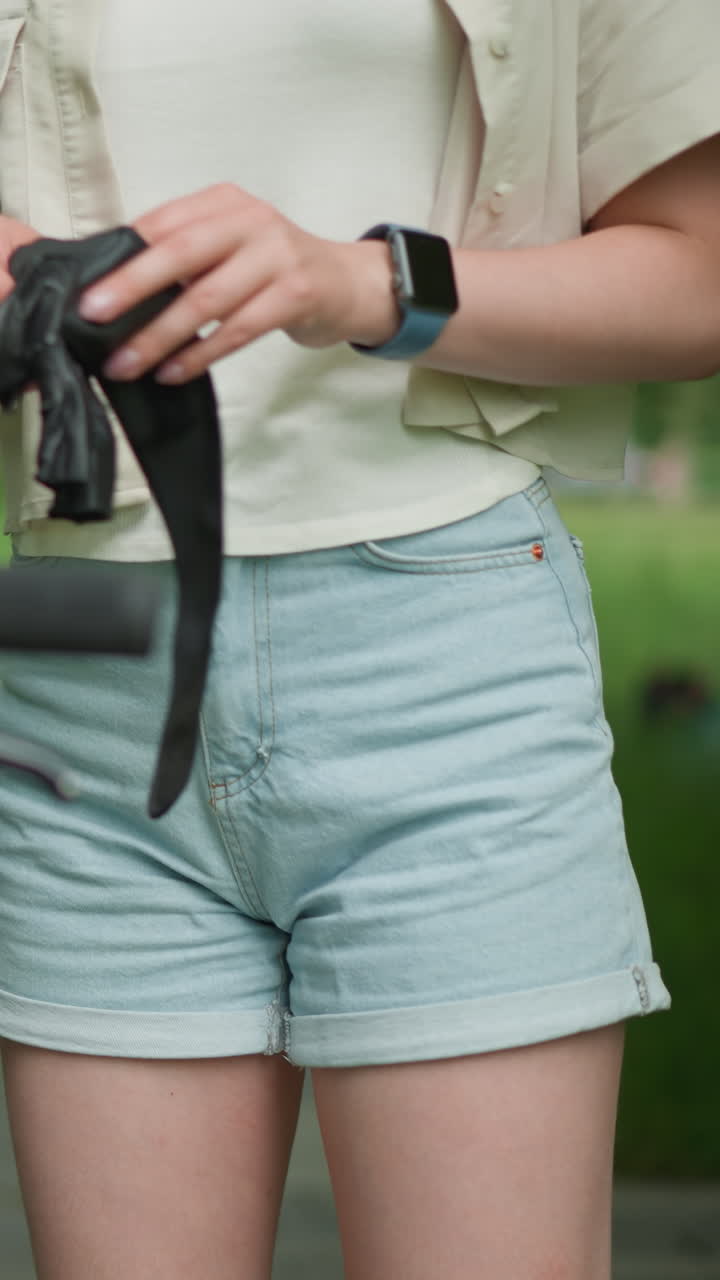 primer plano de una mujer con guantes negros y un reloj inteligente en la mano derecha, preparándose para usar guantes cerca de su bicicleta estacionada en un camino pavimentado, con vegetación borrosa y edificio en el fondo