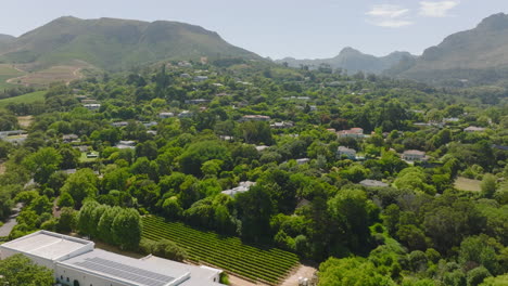 Fly-above-village-or-town-borough-with-residential-houses-surrounded-by-green-trees.-Tilt-up-reveal-of-mountains-in-background.-Cape-Town,-South-Africa