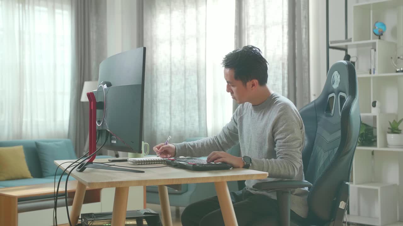 Asian Man In Long Sleeved T-Shirt And Black Pants Looking At Desktop Computer Screen And Writing In Notebook On A Table While Working At Home.