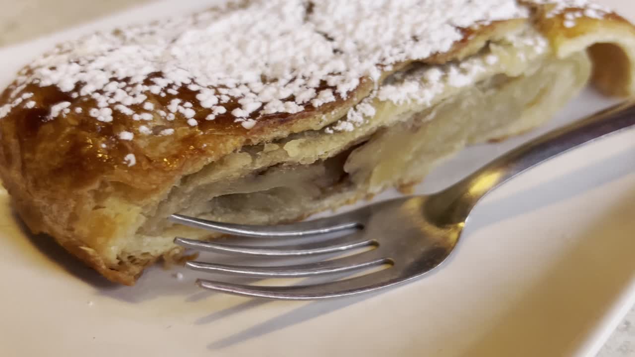 Close-up of a fork near a slice of apple strudel generously dusted with powdered sugar, ready to be enjoyed, showcasing the flaky pastry and sweet filling