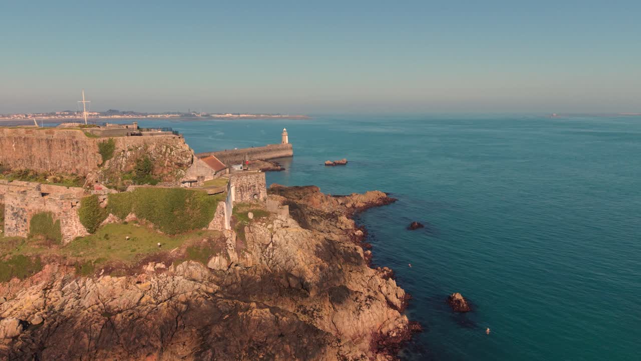 Castello Cornet, St Peter Port, Guernsey. Riprese a bassa quota con drone sopra il castello verso il faro nell'ora d'oro con cielo azzurro, mare limpido, orizzonte velato e viste su Herm e St Sampson oltre.