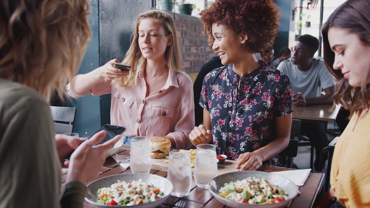 네 명의 여성 친구들이 레스토랑에서 음식 사진을 찍고 소셜 미디어에 올리고 있습니다.