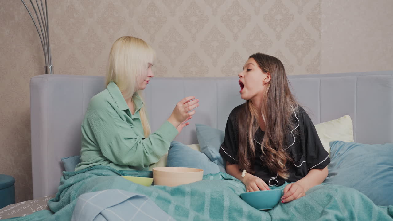 Two girls enjoying playful moment while tossing popcorn into mouths and laughing in cozy indoor setting, lively expressions, comfortable blankets, and shared fun reflect lighthearted bonding