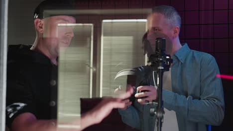 in a red soundproof studio, a man in a blue long-sleeve shirt receives headphones from another man in black. the interaction captures a moment of preparation for a recording session