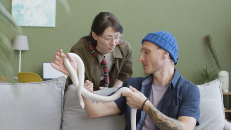 couple holding pet snakes and talking together while the man is sitting and woman is leaning on a comfortable sofa at home 1