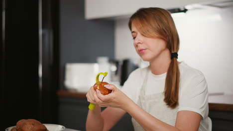 lady in white shirt and ash apron peeling raw potato with yellow peeler at kitchen table, bowl of unpeeled potatoes beside her, background shows blurred kitchen appliances