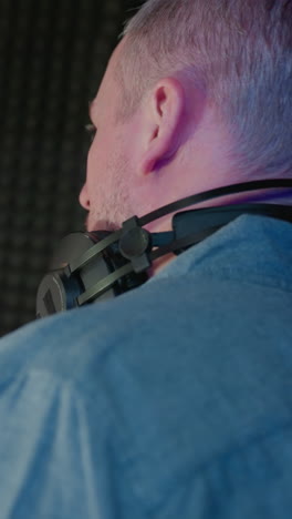 a focused dialogue between two men in a recording studio: one in a black cap and shirt, and the other in a blue shirt with headphones around his neck, against a red and black backdrop