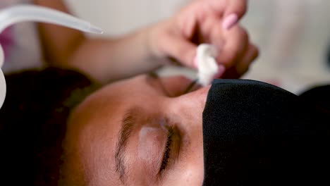 black woman with curly hair is getting her lashes done by an esthetician while at home she wears a mask to protect from covid 19