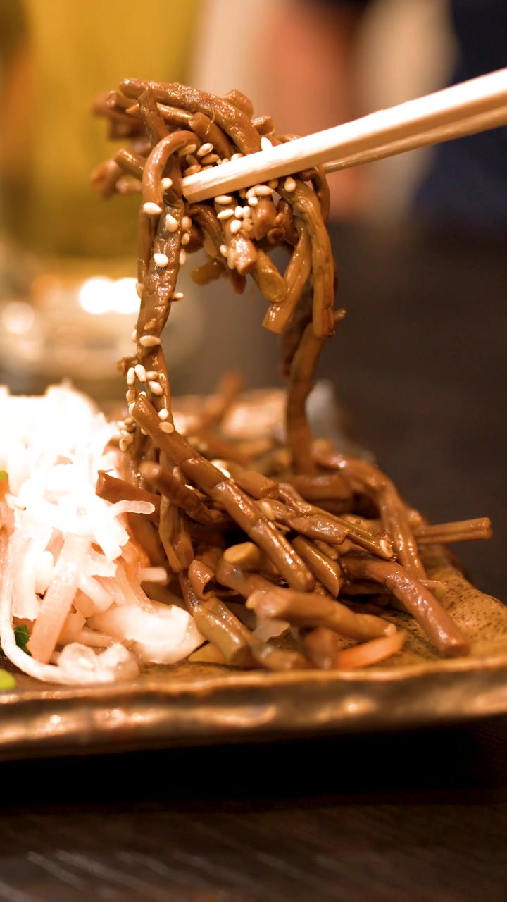 Primo piano di bacchette di legno che sollevano verdure condite da un piatto con illuminazione calda da ristorante.