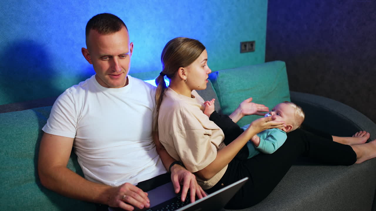 Baby with pacifier lies on mom's laps looking at her intently. Woman leans on her husband working on laptop.