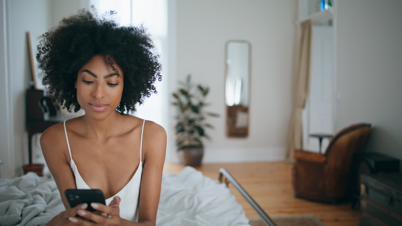 Mulher serena lendo celular na cama em close. Mulher de cabelo cacheado segurando telefone celular.