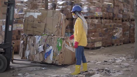 Unloading-of-waste-paper-in-a-warehouse-electric-car-controling-by-female-worker-in-hard-hat-and-yellow-jacket.-Huge-stocks-of-pressed-carton