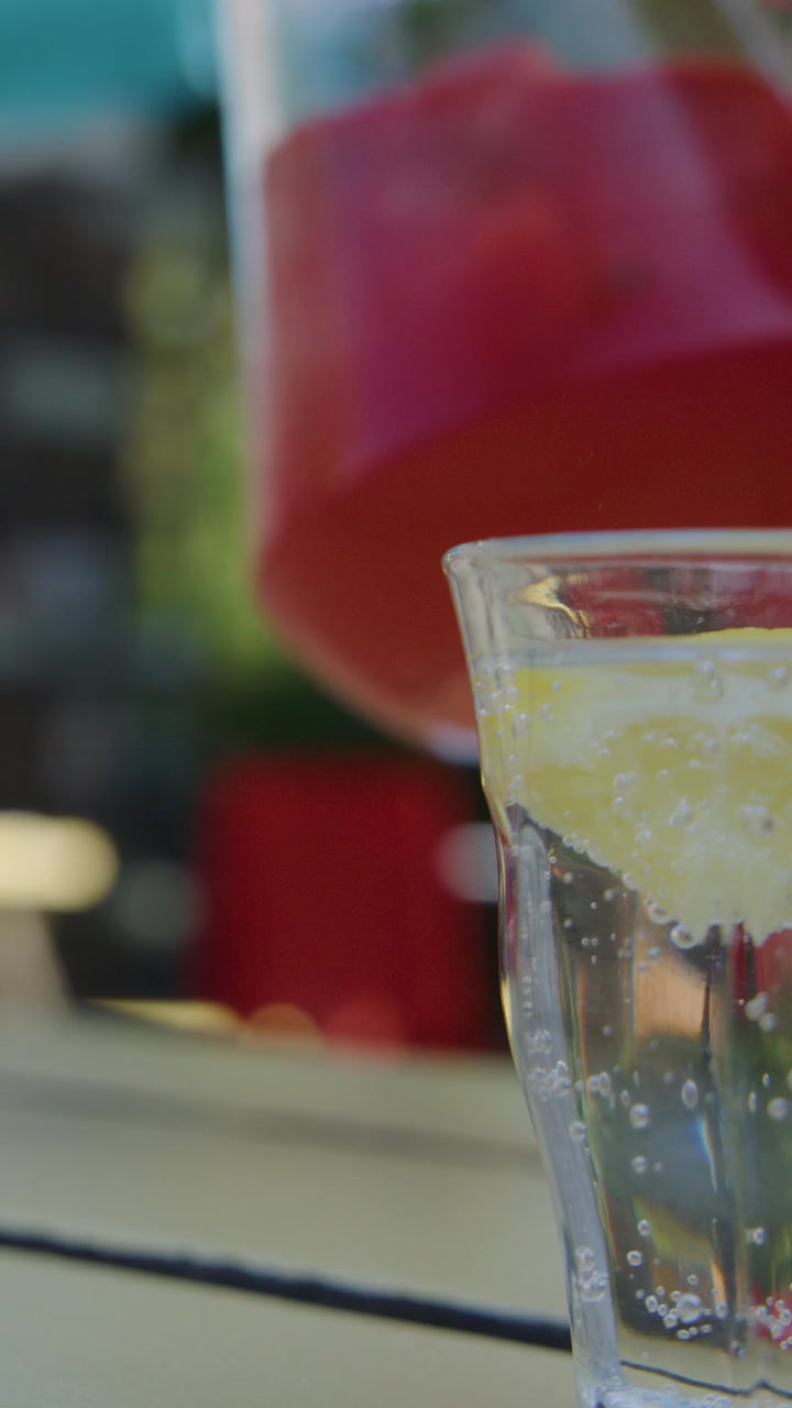 Una mano versa una bevanda rossa da una brocca in un bicchiere di acqua frizzante con limone, in un ambiente esterno soleggiato.