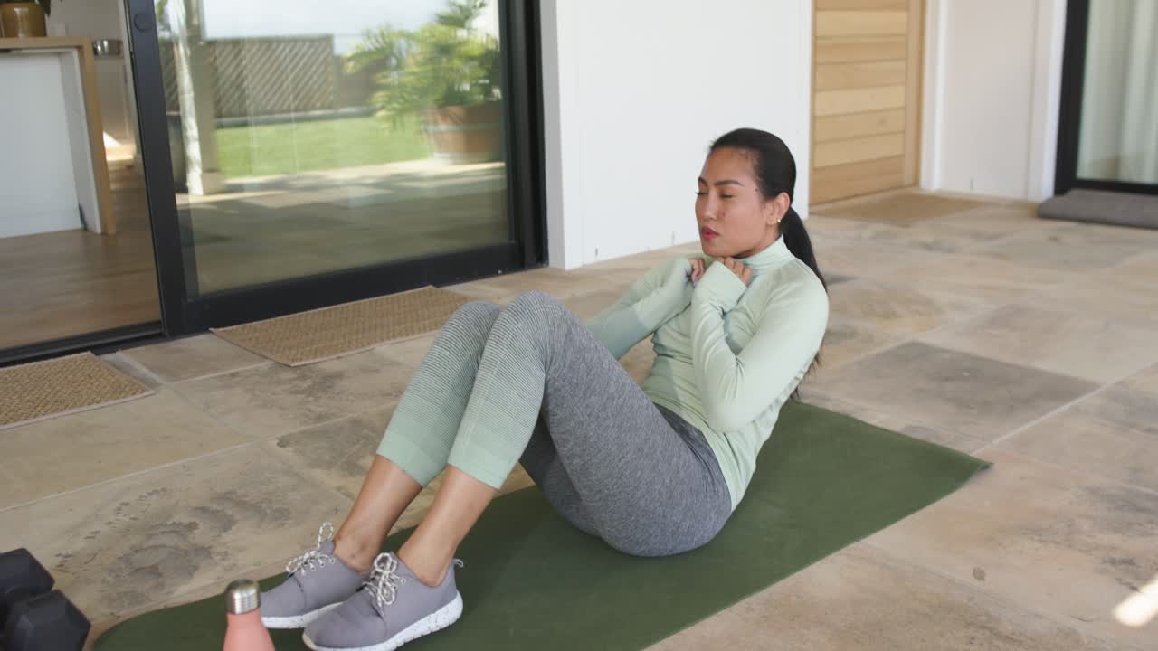 Asian woman clasping chest, crunching on green patio mat by dumbbells and water bottle, copy space