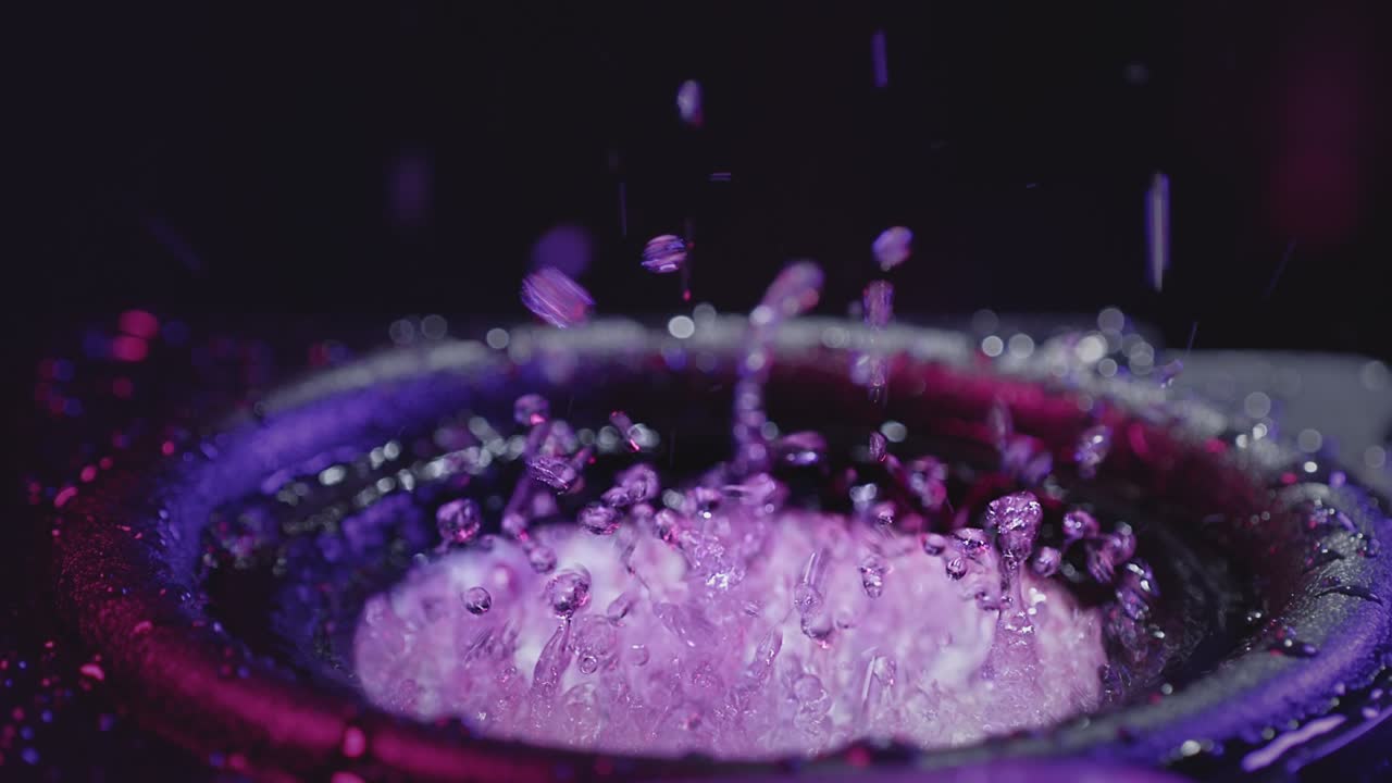 A shaky speaker tosses liquid water in a neon light. Bass sound in a recording studio. Party or concert footage. Pink-blue light on a black background. Retrowave concept.